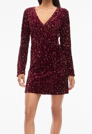 Kobieta ubrana w bordową mini sukienkę z cekinami, długimi rękawami i dekoltem w serek, stojąca na jednolitym białym tle.