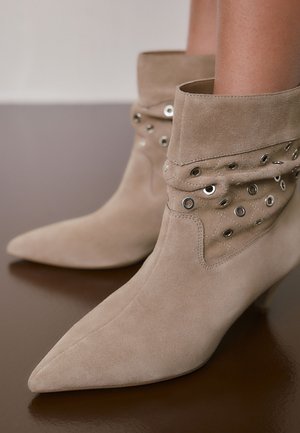 Beige ruskind ankelstøvler med spidse snuder, løstsiddende toppe dekoreret med metaløjer, båret på bare ben mod en brun overflade.