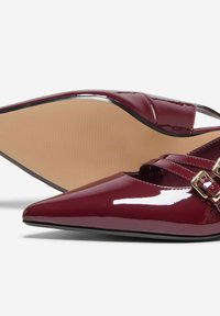 Chaussures pointues en cuir verni bourgogne avec des brides croisées, boucles dorées et une texture lisse ; semelle courbée de couleur marron clair.