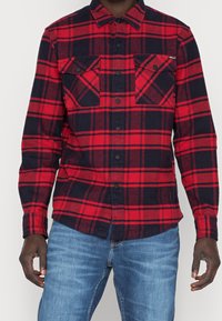 Camicia in flanella a scacchi rossa e blu marina con maniche lunghe, due tasche sul petto, bottoni neri e orlo curvo. Il tessuto sembra morbido e texturizzato.