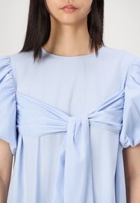 Blouse bleu clair avec des manches bouffantes, présentant un détail noué à l'avant et une texture lisse. Encolure ronde simple, design fluide.