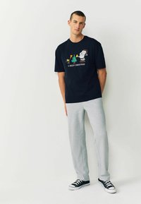 Must musta T-särk, millel on jõuluteemaline graafika, kokku sobitatud hallide dressipükstega. Mudel seisab lihtsate tossudega tasasel taustal.