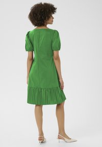 Zelena bombažna obleka z nabranimi rokavi, nabranim plisiranim robom in zadrgo na hrbtu. Dodatki vključujejo svetlo obarvane pete.