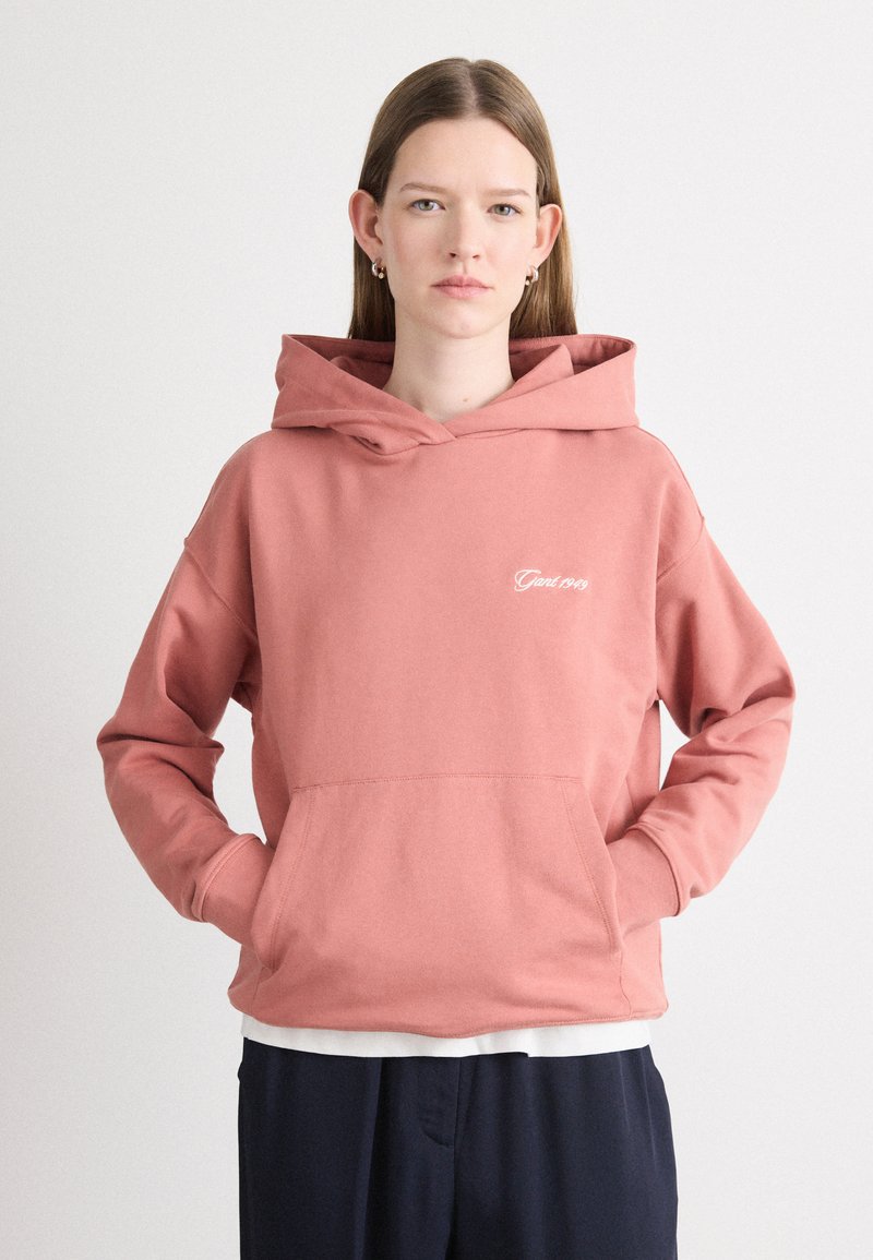 Różowy hoodie z przednią kieszenią i kapturem na sznurek, z białym napisem na klatce piersiowej. Miękka tkanina o luźnym kroju.