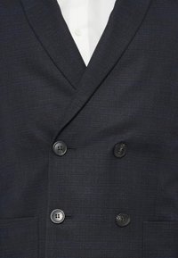 Blazer azul marino de doble botonadura con una tela texturizada, que presenta dos botones negros y un bolsillo en el pecho, combinado con una camisa blanca debajo.