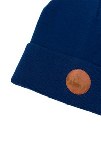 Niebieska dziana czapka z podwiniętym mankietem, z okrągłą brązową skórzaną łatą wytłoczoną z motywami gór i drzew.