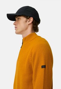 Oranger, gerippter Strickpullover mit hohem Kragen und schwarzem Reißverschluss, kombiniert mit einer schwarzen Mütze. Verfügt über ein Markenlogo auf dem Ärmel des Pullovers.