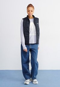 Zwarte gewatteerde vest over een lichtgrijs langharig shirt, gecombineerd met losse blauwe jeans en witte sneakers. Eenvoudige achtergrond.