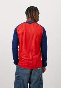 Man med flätat hår som bär en röd och marinblå långärmad tröja med texten "España" på bakre kragen och lösa blå denim cargobyxor.