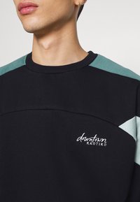 Sudadera negra con acentos en color verde azulado en los hombros y texto bordado en blanco que dice "downtown KAOTIKO", con cuello redondo y una textura suave.