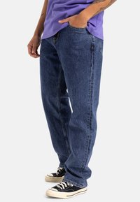 Vaqueros de denim azul con corte recto, que cuentan con cinco bolsillos, detalles de costura sutiles y se combinan con zapatillas negras de corte bajo.