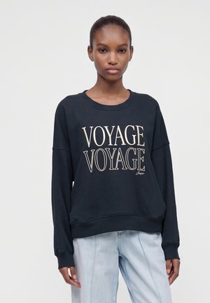 Млада жена с тъмна коса, облечена в червена блуза с надпис "VOYAGE VOYAGE" и светлосини дънки на едноцветен светъл фон.