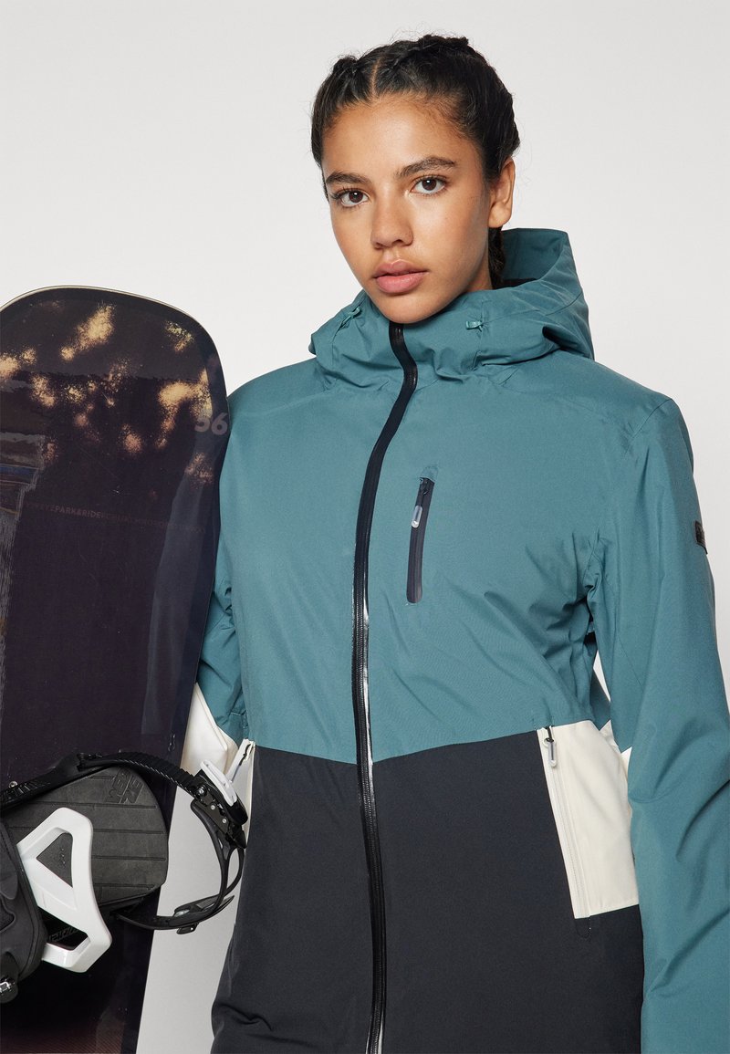 Wasserdichte Skijacke in Teal, Schwarz und Creme. Verfügt über eine mit Reißverschluss verschlossene Brusttasche, eine verstellbare Kapuze und eine glatte, matte Textur. Im Hintergrund ein Snowboard.