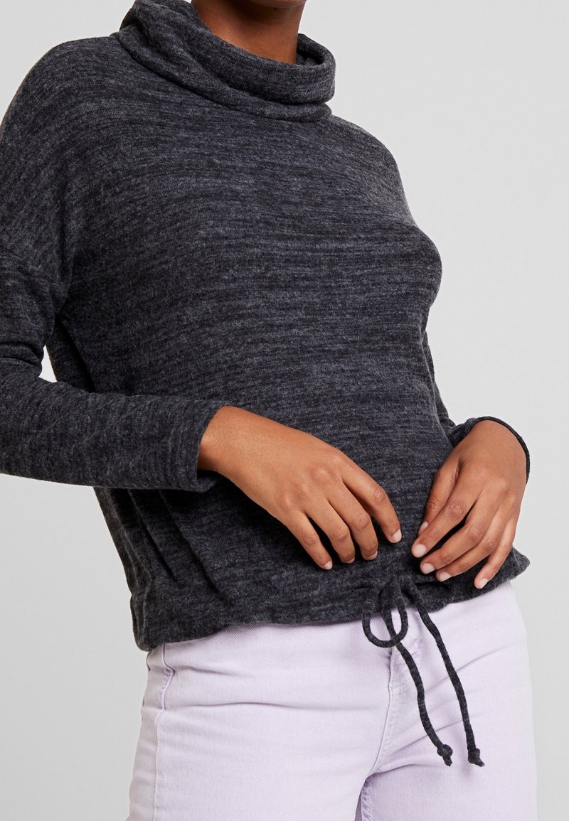 Pull à col roulé gris, texturé, avec un ourlet à cordon de serrage. Manches longues et coupe décontractée, associé à un pantalon violet clair.