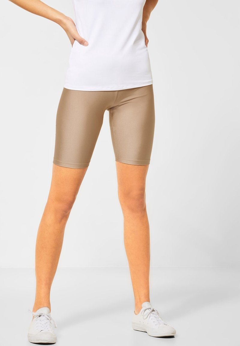 Beżowe rowerowe szorty z wysokim stanem, o gładkiej fakturze, dopasowanym kroju, kończące się w połowie uda, w zestawieniu z białą bluzką i białymi trampkami.