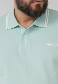 Hellgrün mintfarbenes Polohemd mit strukturiertem Gewebe, klassischem Kragen mit weißen Streifen, zwei Knöpfen und einem kleinen Logo auf der Brust.