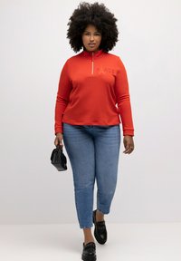 Sudadera roja con cremallera y texto en relieve "PARÍS", combinada con jeans de color azul claro y mocasines negros. El modelo sostiene un pequeño bolso negro.