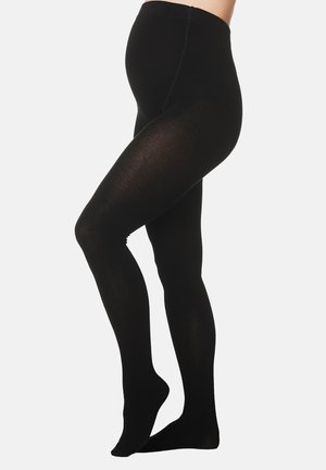 Schwangere Person trägt blickdichte schwarze Umstandstrumpfhosen und steht auf Zehenspitzen vor weißem Hintergrund.