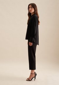 Czarna bluzka z długim rękawem o luźnym kroju i mankietami zapinanymi na guziki, zestawiona z dopasowanymi czarnymi spodniami i czarnymi sandałami na wysokim obcasie.