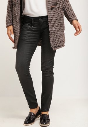 Persoon die zwarte slim-fit broek draagt, zwarte loafers met franjes, een wit topje en een grof gebreid taupekleurig vest met toggles.