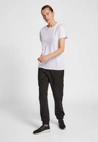 Biała koszulka z krótkim rękawem w połączeniu z czarnymi spodniami dresowymi. Model nosi czarne buty sportowe. Gładki materiał, luźny krój, styl casual.