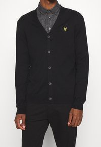 Cardigan noir en maille, doté d'un col en V, d'une fermeture à boutons et d'un petit logo d'aigle jaune sur la poitrine gauche.