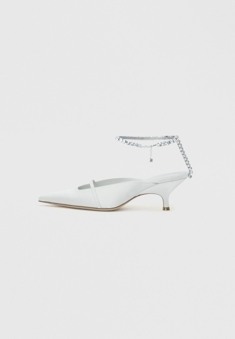 Mule branco de salto agulha baixo com biqueira pontiaguda e detalhe de tira para o tornozelo com strass, sobre um fundo liso.