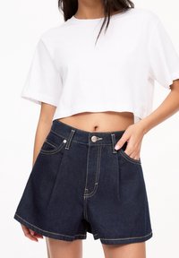 Camiseta blanca recortada de manga corta combinada con pantalones cortos de mezclilla oscuros de tiro alto que presentan pliegues en la parte delantera y costuras en contraste.