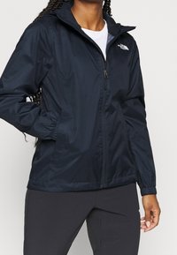 Laivastonsininen tuulta estävä takki, jossa on vetoketjulla varustettu etuosa, joustavat hihansuut ja huppu. Oikealla rintapoiminnalla on The North Face -logo, ja se on valmistettu kevyestä materiaalista.