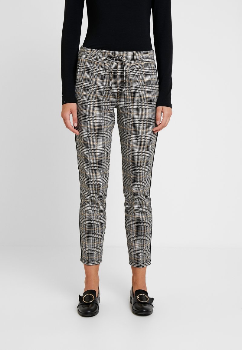 Femme portant un pantalon slim à carreaux avec taille à cordon, associé à des mocassins noirs avec des détails de boucle dorée.