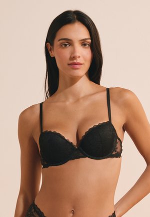 Woman with long dark hair wearing a black lace bra against a neutral beige background.