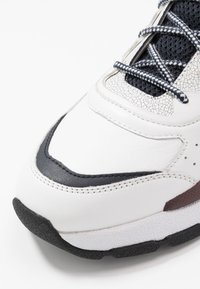 Sneaker bianco con pelle testurizzata, suola in gomma nera e pannellatura in mesh scuro. Presenta cuciture intricate e lacci a fantasia.