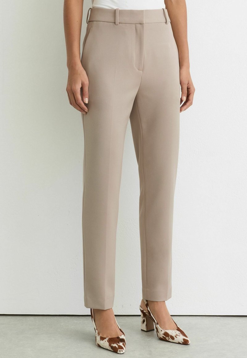 Femeie purtând pantaloni bej croiți și pantofi cu toc înalt, maro cu alb, stând în fața unui perete alb simplu.