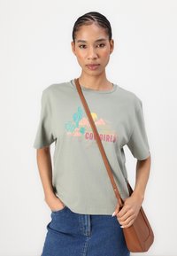 Light green cotton t-shirt with short sleeves and "Cowgirls" graphic. Paired with a denim skirt and a brown crossbody bag.