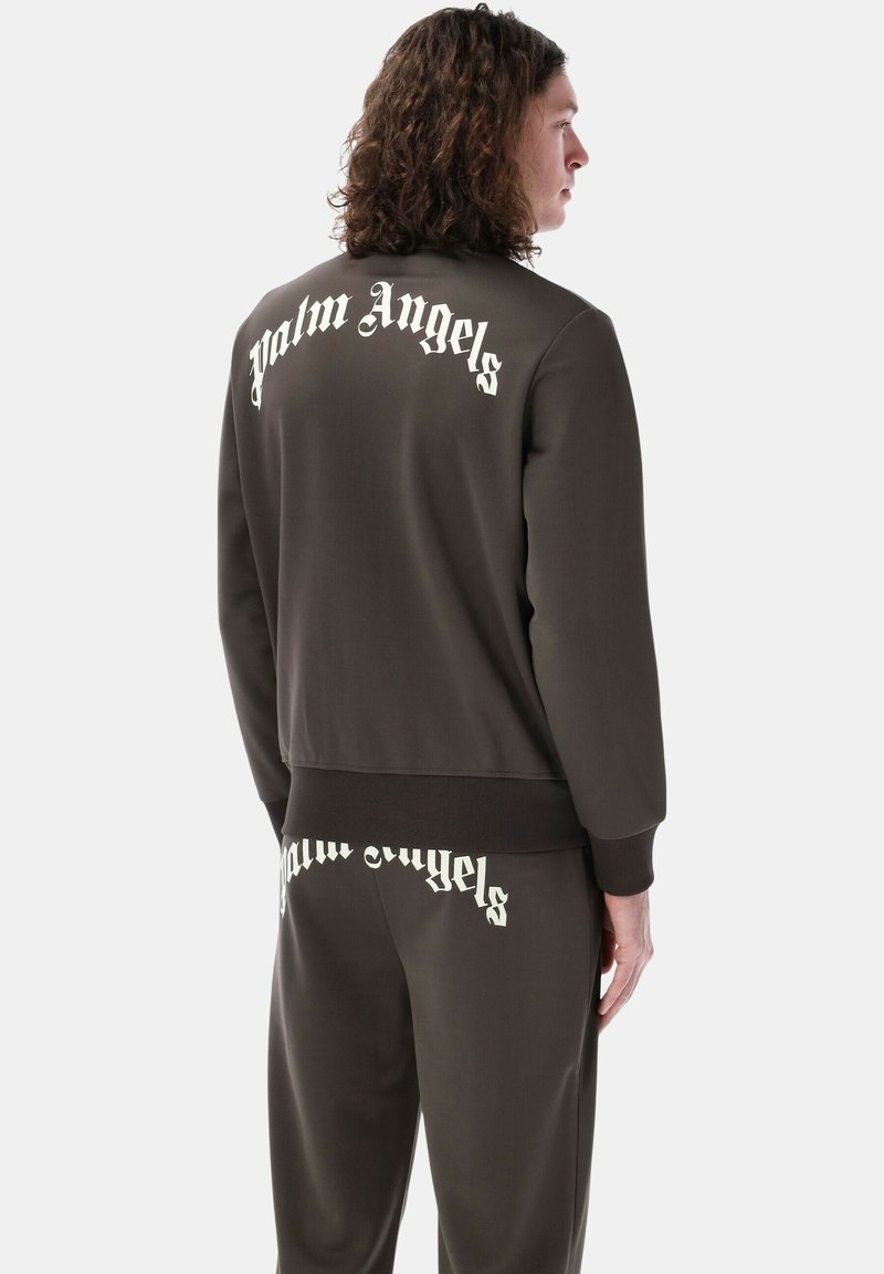 Tuta di colore marrone scuro composta da una giacca con zip e pantaloni con vita elastica. Entrambi presentano il logo "Palm Angels" in bianco sul retro.