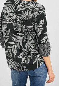 Blusa nera con motivo a foglie bianche, maniche lunghe con dettagli floreali e vestibilità ampia. Il design sul retro include un dettaglio plissettato.