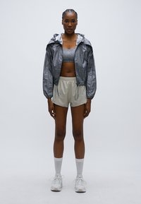 Γκρι σακάκι με κουκούλα και ελαφρύ γυαλιστερό φινίρισμα, αθλητικό μπουστάκι σε ίδιο χρώμα και μπεζ σορτς. Το μοντέλο φοράει λευκές κάλτσες και αθλητικά παπούτσια, στέκεται μπροστά σε ένα απλό φόντο.