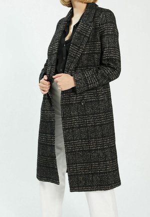 Femme vêtue d'un long manteau à carreaux noirs et gris par-dessus un chemisier noir et un pantalon blanc, tenant les revers du manteau des deux mains.