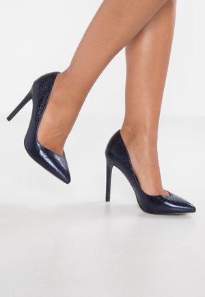 Marineblauwe hoge hakken met een puntige neus, glanzende textuur en een slank ontwerp. Voorzien van een slanke stilettohak voor extra hoogte.