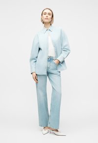 Γυναίκα που φορά γαλάζιο τζιν πουκάμισο και τζιν παντελόνι, λευκή μπλούζα, λευκά τακούνια, στέκεται με ένα χέρι στην τσέπη μπροστά σε λευκό φόντο.