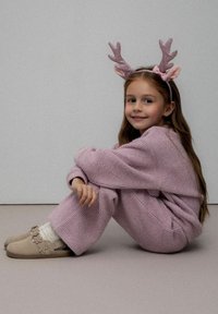 Młoda dziewczynka siedząca z nogami skrzyżowanymi, ubrana w różowy dziergany komplecik, beżowe kapcie i świecącą różową opaskę z porożem renifera.
