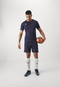 Tamsiai mėlyna sportinė apranga, kurioje yra marškiniai trumpomis rankovėmis, šortai, balti kojinės ir spalvingi futbolo batai, laikantis futbolo kamuolį.