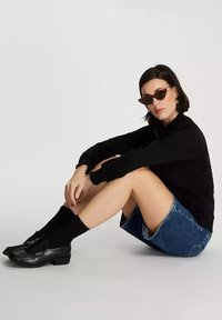 Czarny sweter, dżinsowa spódnica, czarne skarpetki i czarne loafersy. Brązowe okulary w kształcie kociego oka. Model w siedzącej, zrelaksowanej pozycji na jednolitym tle.