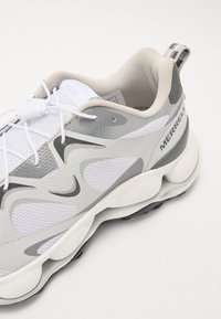 Chaussure de sport blanche avec un design texturé, présentant des accents gris et une tige en mesh. Des lacets ronds et une semelle rembourrée sont visibles.