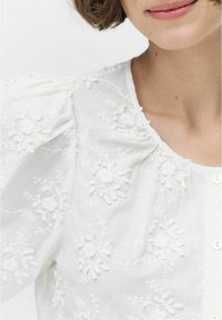 Witte blouse met bloemenborduursels en verhoogde texturen, een ronde halslijn, en korte pofmouwen, vastgemaakt met kleine knopen.