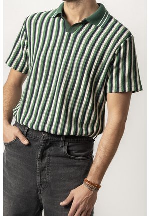 Homme portant un polo en maille rayé vert, blanc et marine à manches courtes, jean noir et bracelets au poignet droit.