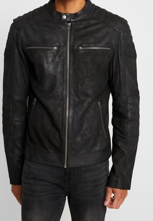 Homme portant une veste en cuir noire cintrée avec des poches zippées sur la poitrine et les côtés, et un col à boutons-pression.