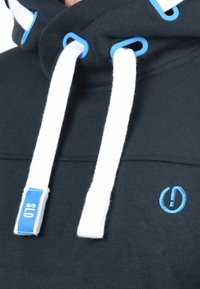 Donkerblauwe hoodie met witte trekkoorden en lichtblauwe hardware. Heeft een klein blauw logo en een zachte stoftextuur.