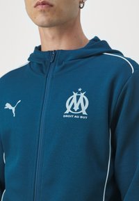 Puma džemperis su užtrauktuku žaliai mėlynos spalvos audinyje, su balto žvaigždės logotipu ir tekstu "DROIT AU BUT", su kontrastingomis baltomis šoninėmis juostomis.