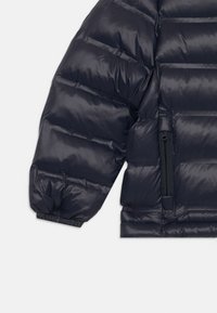 Puffjacka i marinblått, tillverkad av glänsande nylon. Har quiltad design, elastiska muddar och en dragkedjeficka på sidan.
